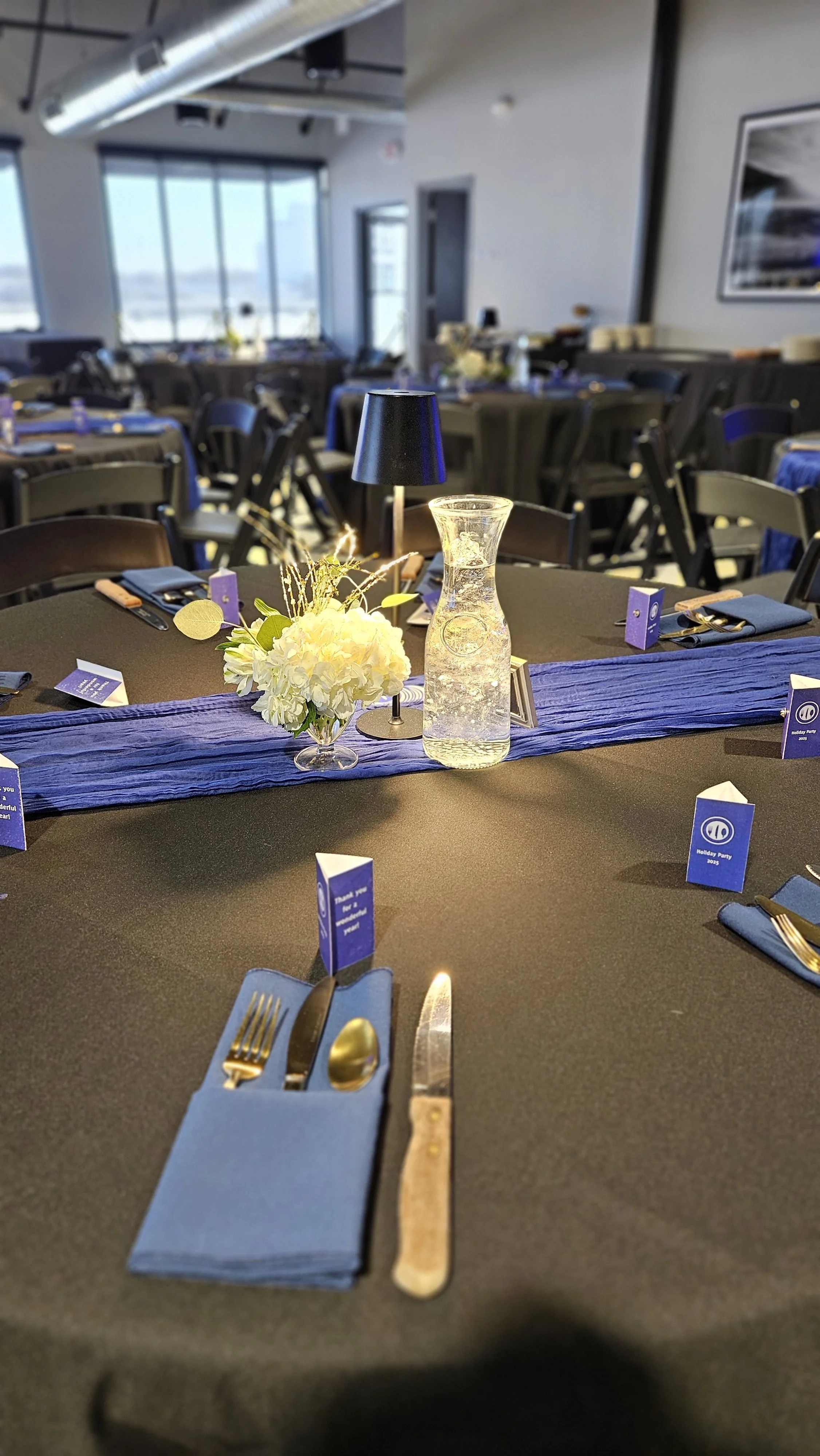 Decorated banquet table set with blue napkins, gold utensils, a small black lamp, a blue flower arrangement, a glass water pitcher, and small purple place cards in a well-lit modern event space.