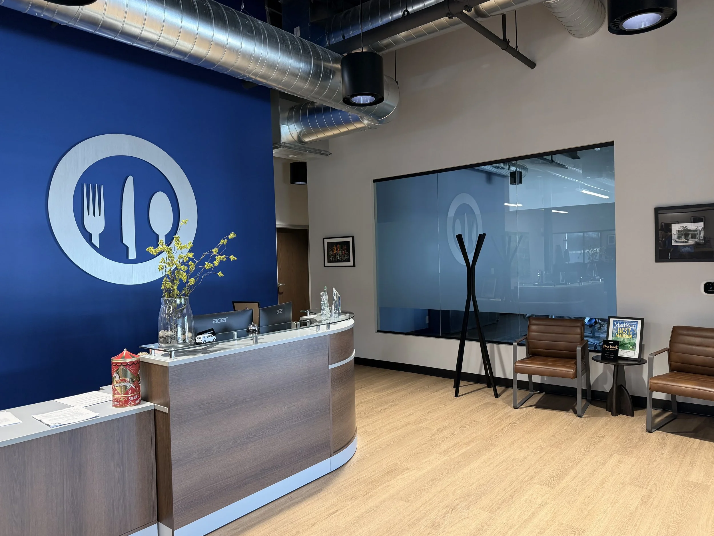 Hotel reception area with a blue accent wall, large utensils logo, and a curved front desk with vases and computers. Seating area with two brown chairs, a small table with magazines, and a large mirror or window reflecting the room.