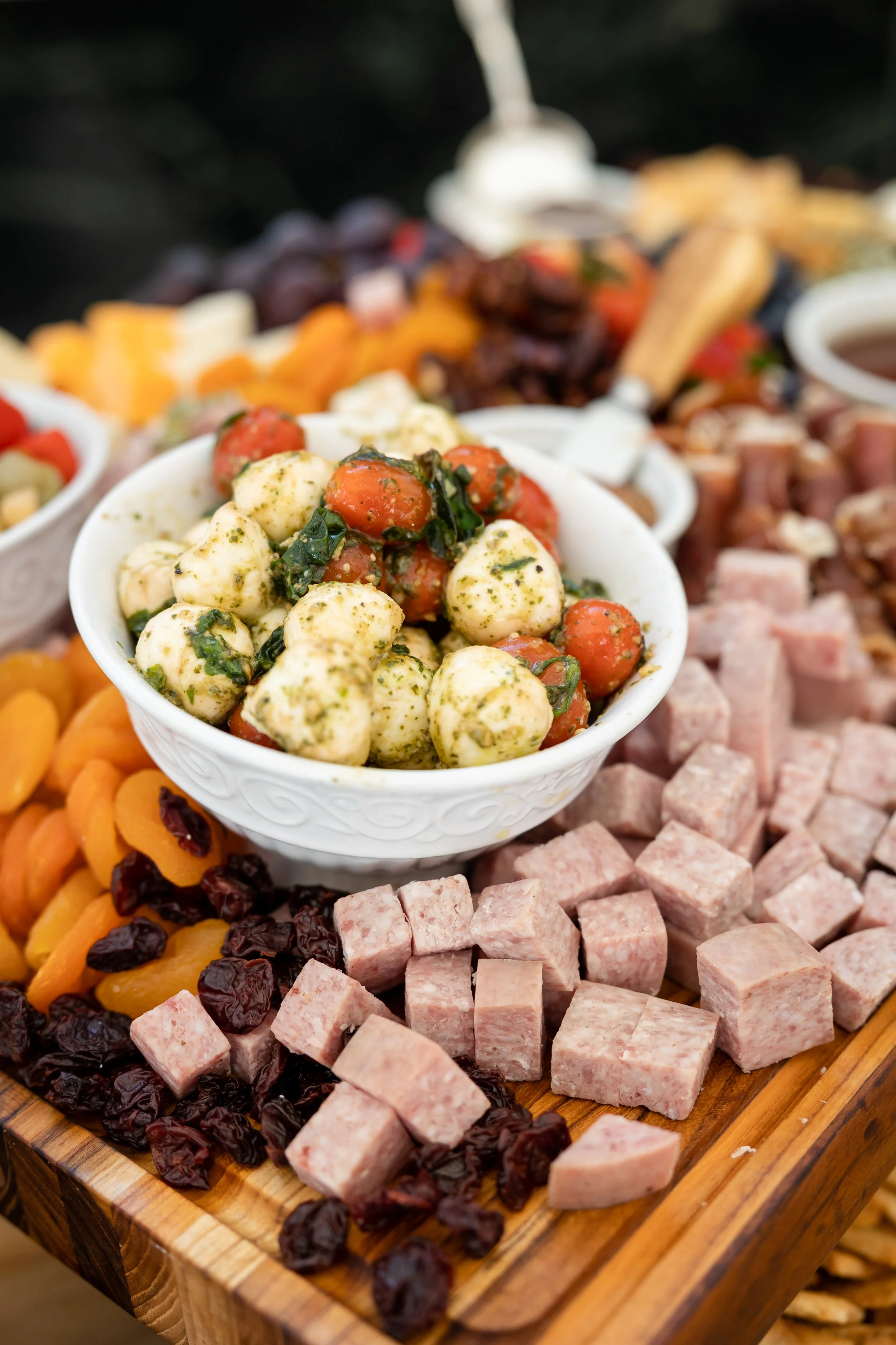 A bowl of marinated mozzarella balls and cherry tomatoes on a wooden serving board, surrounded by diced ham, dried cranberries, purple dried apricots, and other snack items.