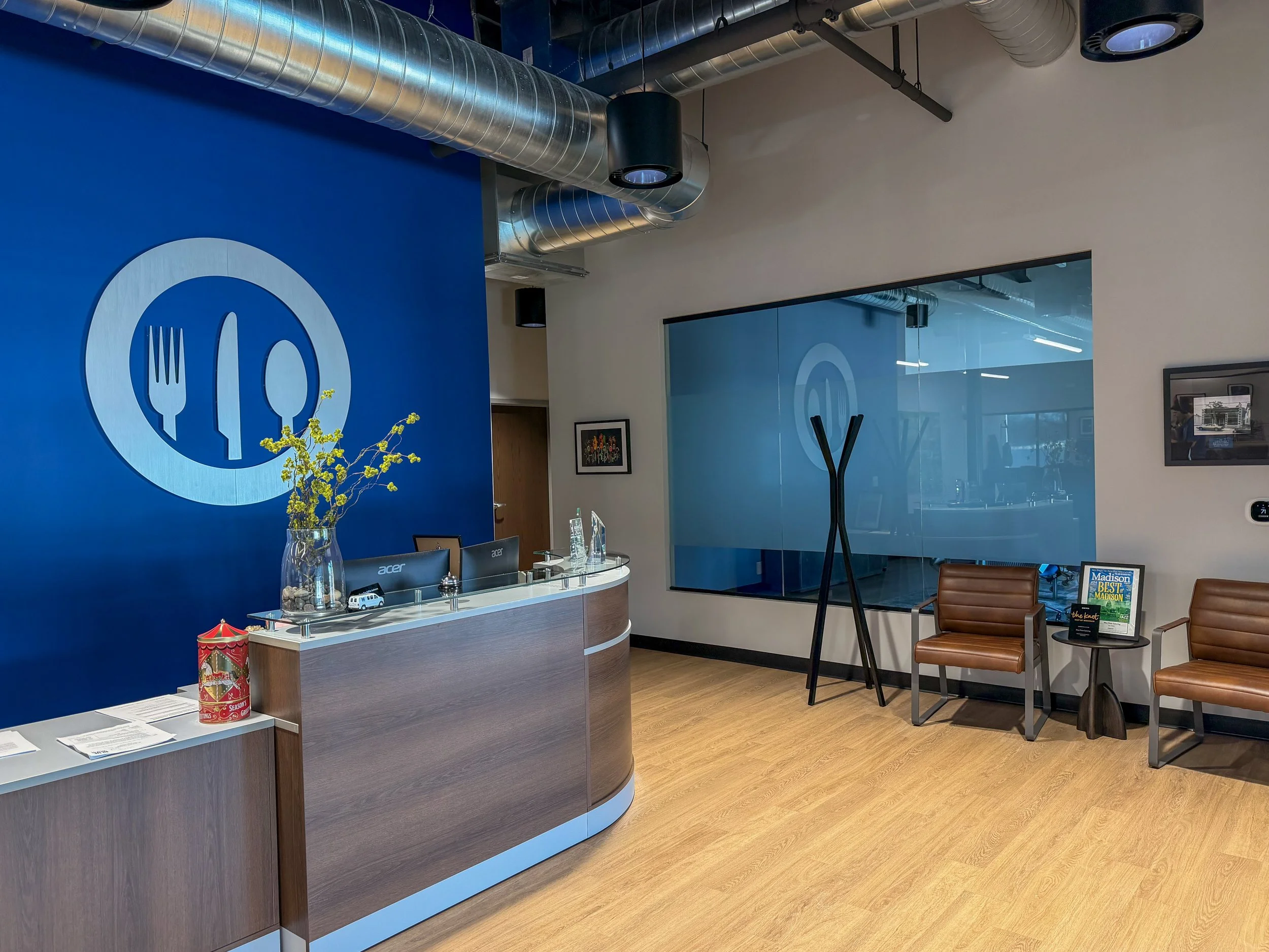 Hotel reception area with a blue accent wall displaying a fork, knife, and spoon logo. There is a curved front desk with computers, a large glass vase with yellow flowers, and a red decorative container. To the right, there are two brown chairs with 