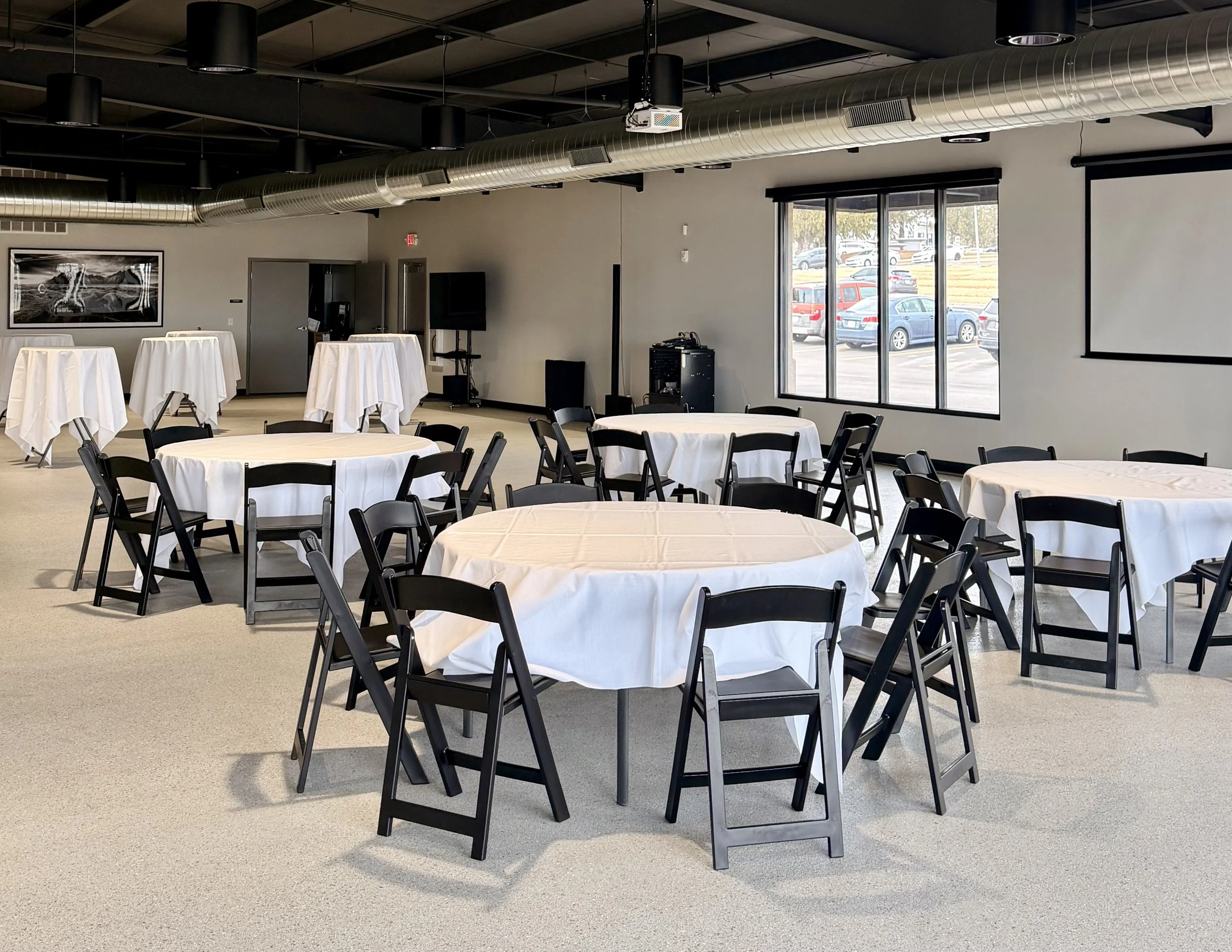 Event space with round tables covered in white tablecloths and black chairs, large windows, and ceiling-mounted ventilation ducts.