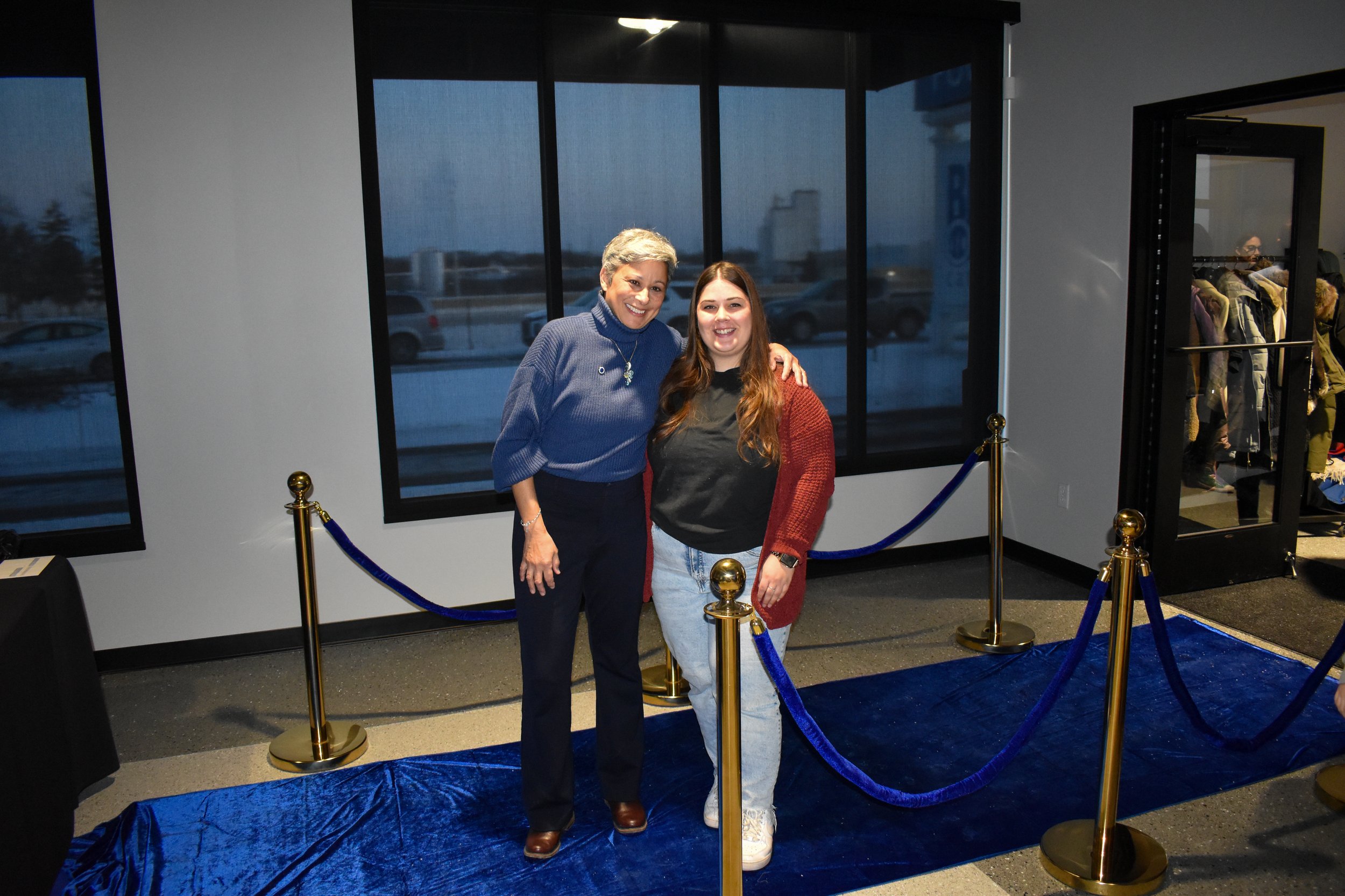 Two women standing close together, smiling, against a backdrop of large windows and a blue velvet rope barrier on a blue carpet in an indoor space.