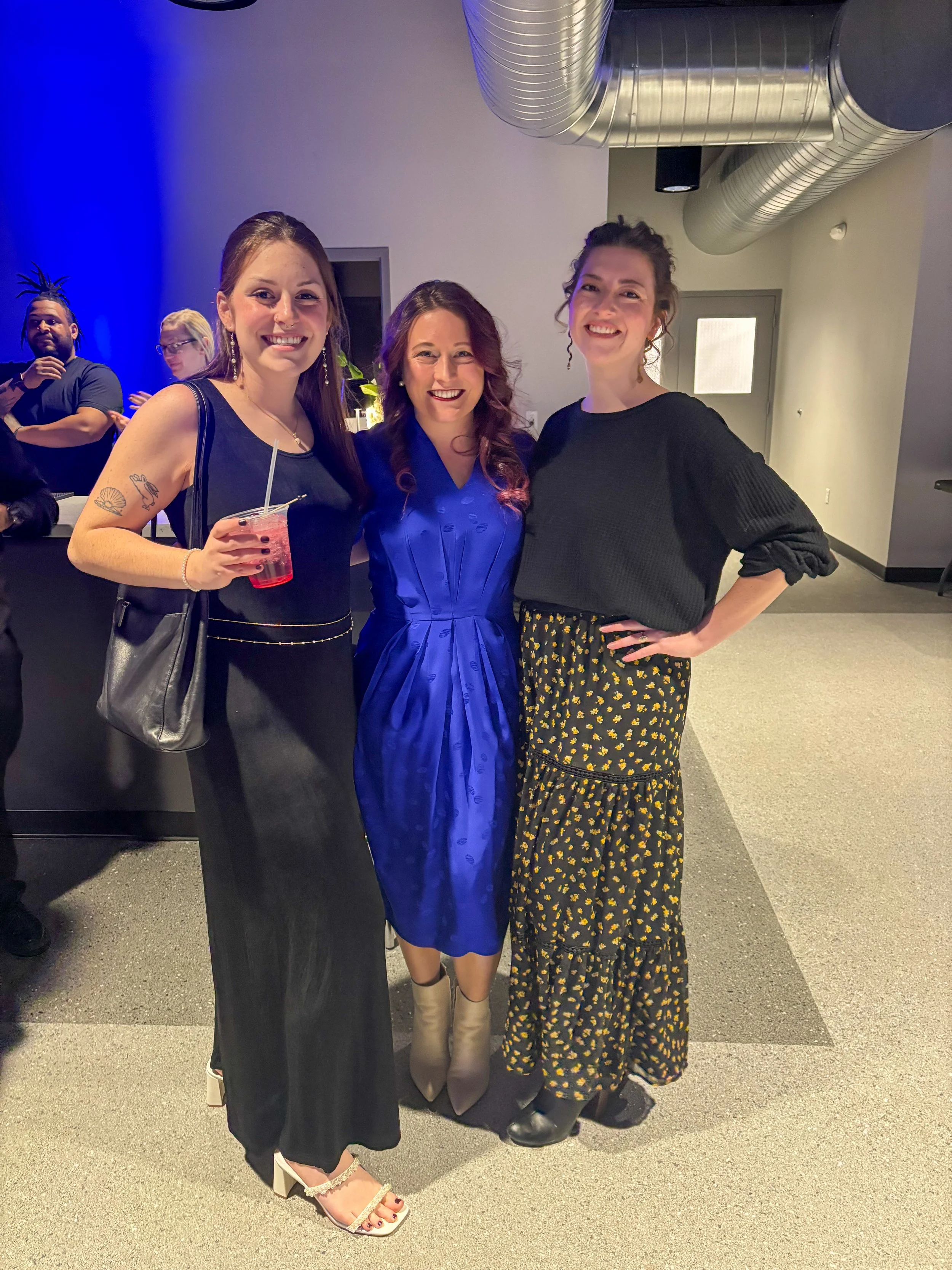 Three women standing together at an indoor event, smiling for the photo. The woman on the left is holding a pink drink, the woman in the middle is wearing a blue dress, and the woman on the right is wearing a black top and a floral skirt.