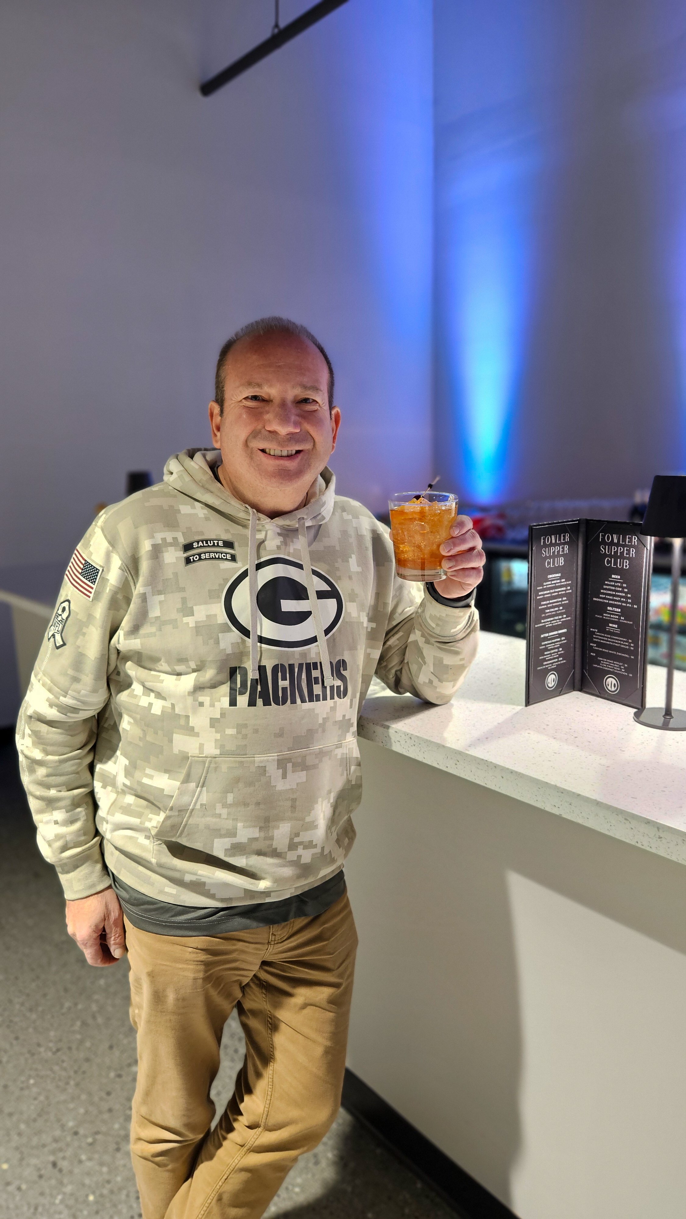 A smiling man in Green Bay Packers hoodie holding a drink at a bar or lounge.