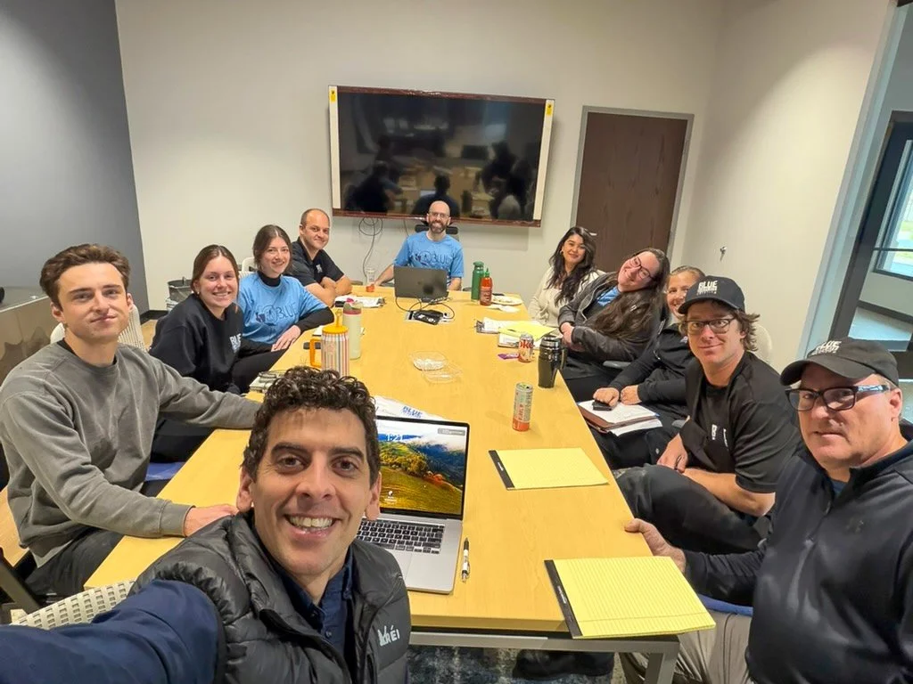 A group of fifteen people sitting around a large conference table in a meeting room, smiling at the camera. There are laptops, notebooks, and drinks on the table, with a large screen on the wall behind them.