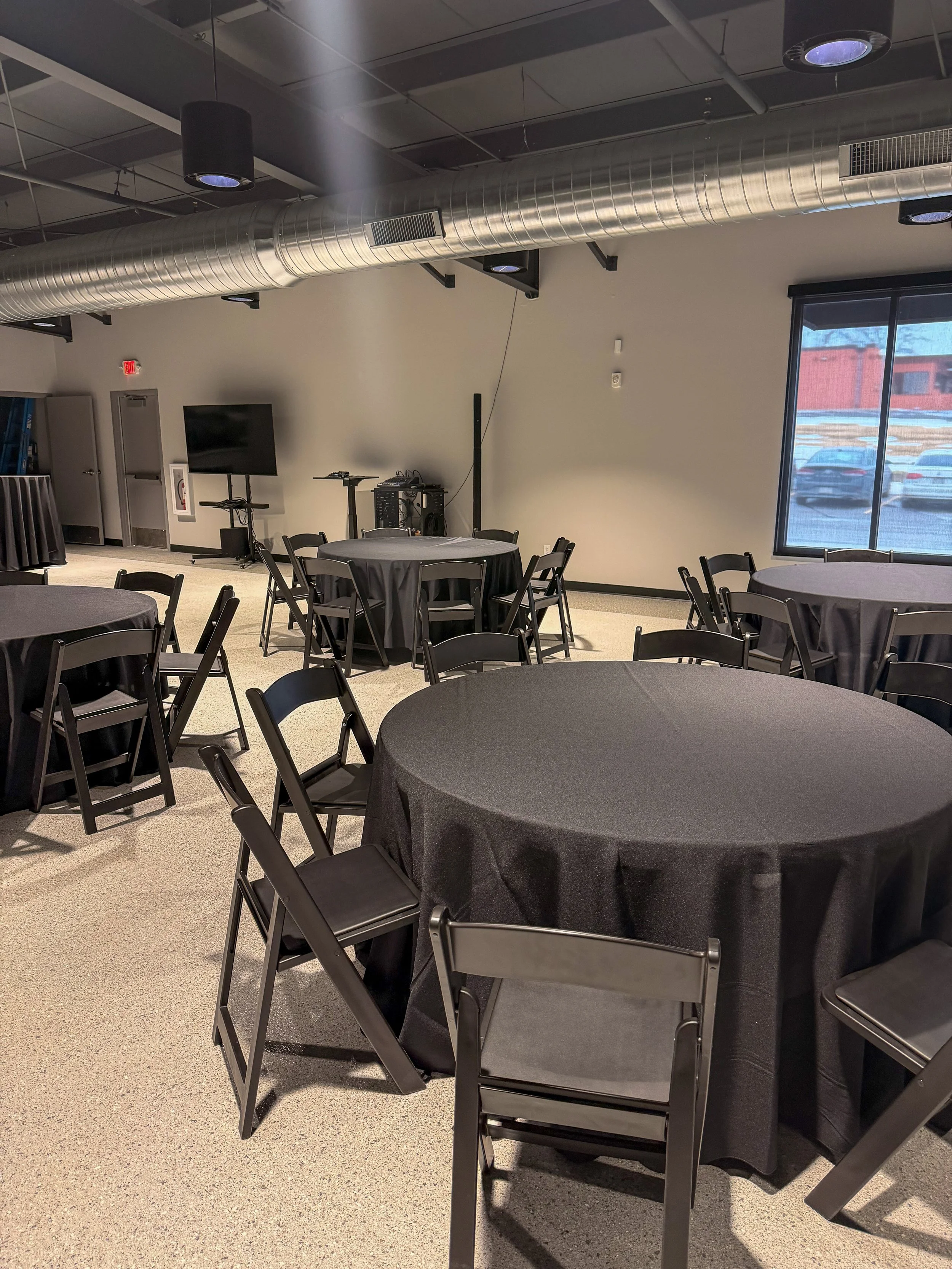 Empty banquet hall with round tables covered in black tablecloths and black folding chairs, featuring a large window on the right and a flat-screen TV on a stand in the background.