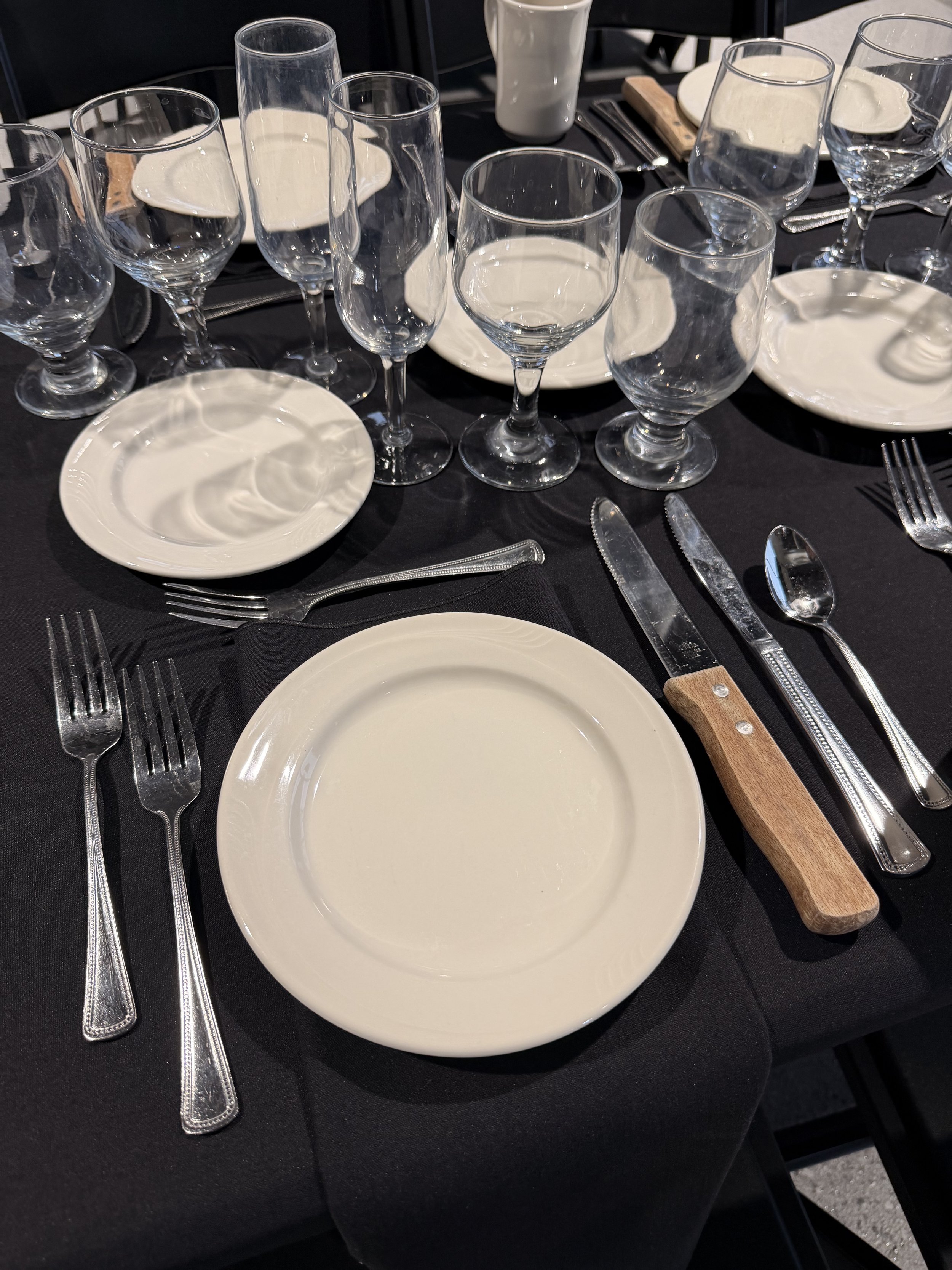 A formal dining table set with white plates, silver cutlery, several wine glasses, and water glasses on a black tablecloth.