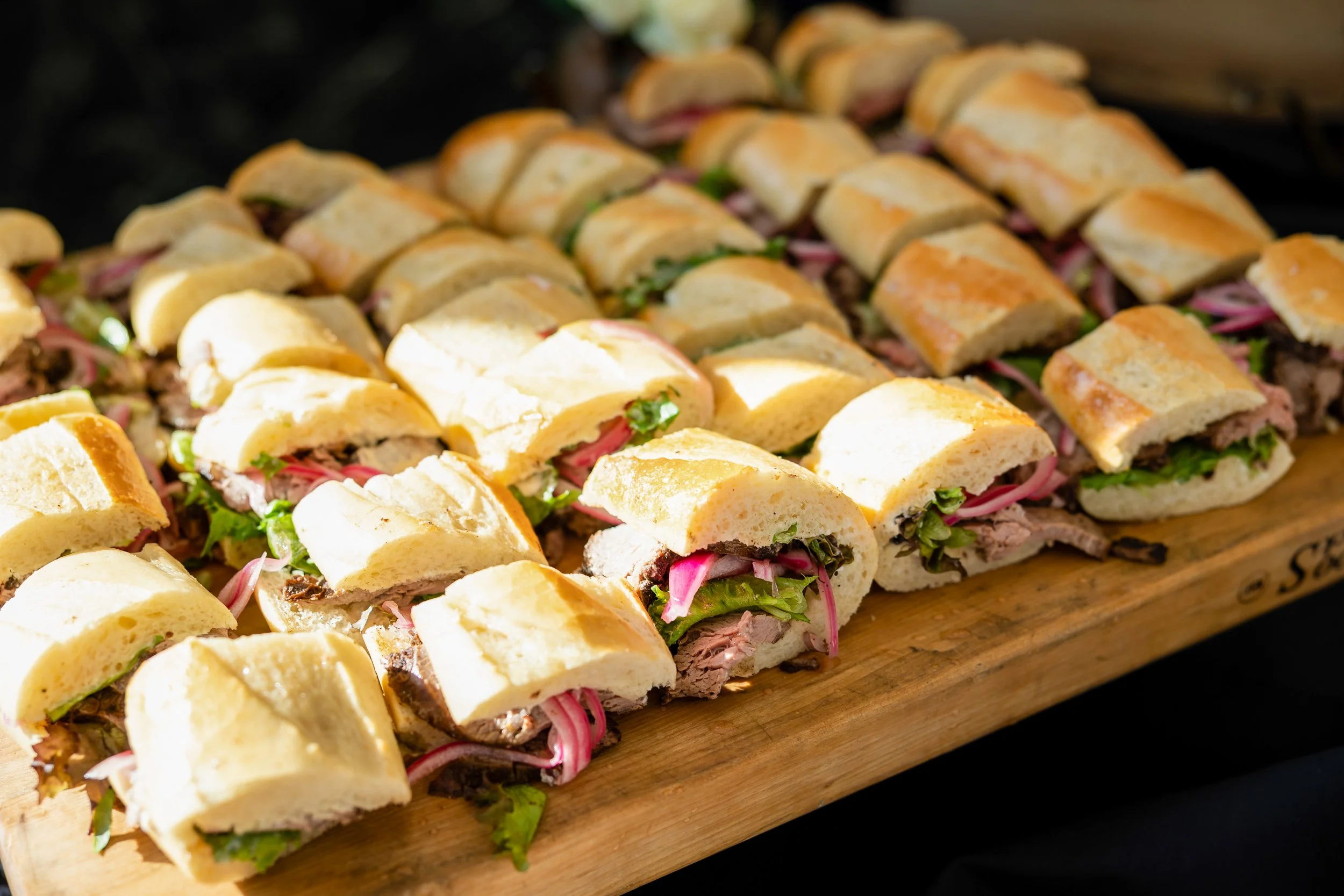 Tray of sliced sandwiches with meats, lettuce, and pickled onions on a wooden platter.