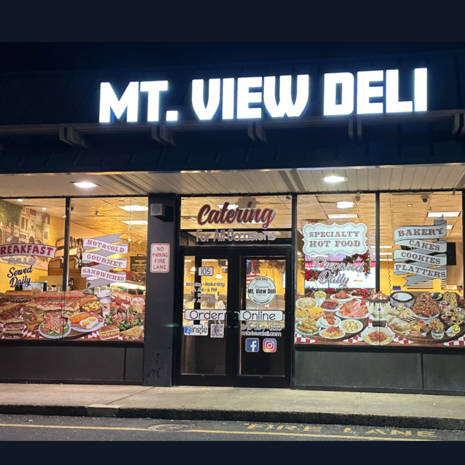 Exterior view of MT. VIEW DELI at night, with bright signage, large display windows showcasing breakfast and catering options, and various signs advertising food items like bakery, hot food, sandwiches, and cookies.