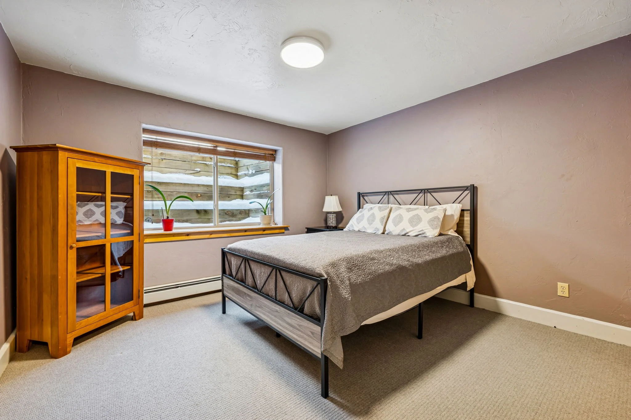 A bedroom with beige walls, a bed with a black metal frame and light gray bedding, a wooden cabinet with glass doors, a window with wooden blinds, two potted plants on the windowsill, a small nightstand with a lamp, and a beige carpeted floor.