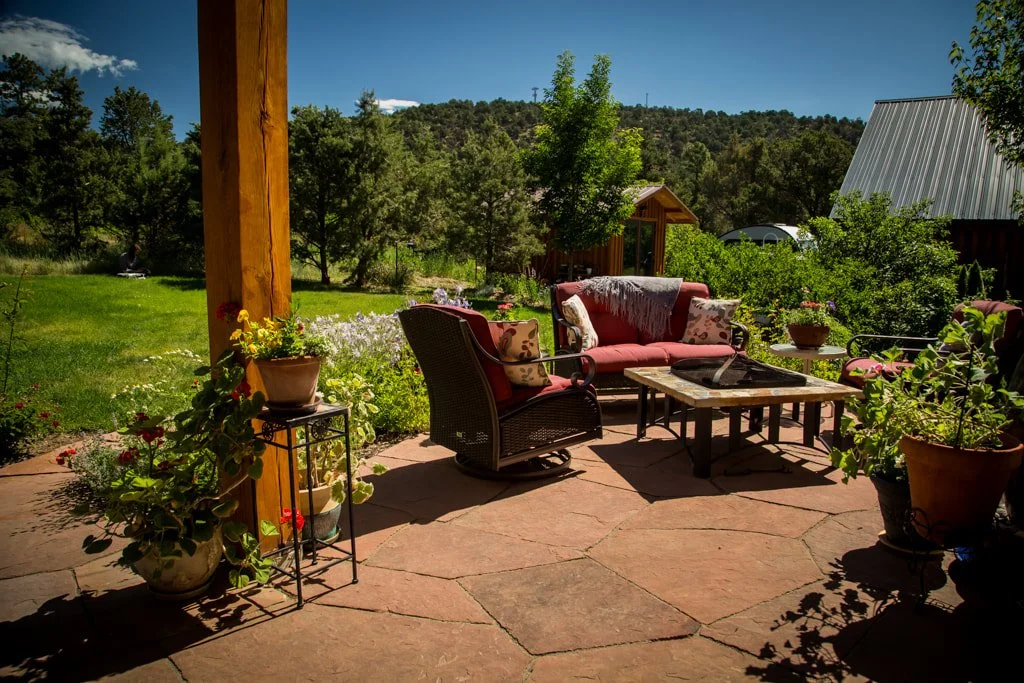 Outdoor patio with wicker and cushioned chairs, a wooden coffee table, and potted plants, overlooking a grassy yard with trees and a mountain landscape in the distance.