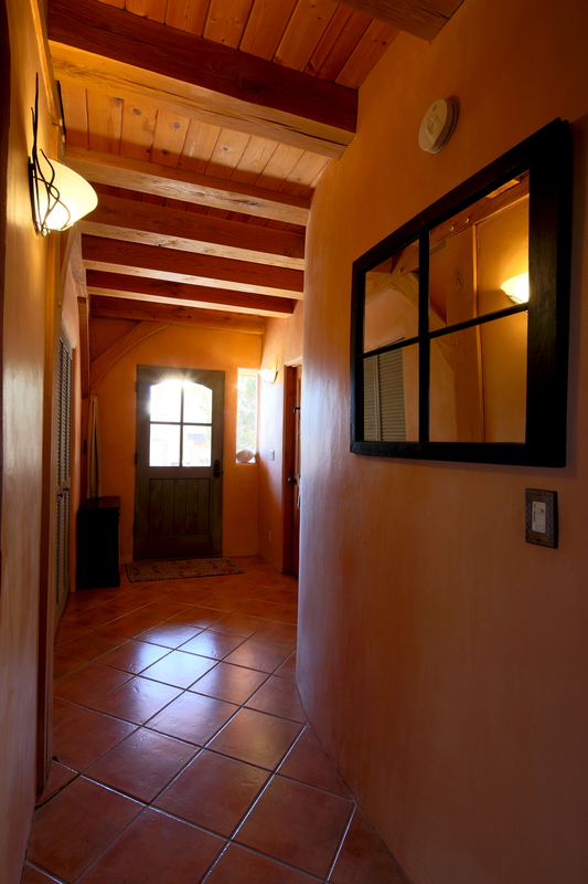Indoor hallway with terracotta tile flooring, wood-beamed ceiling, and peach-colored walls. A windowed front door with glass panels, a mirror on the right wall, and a smoke detector are visible.