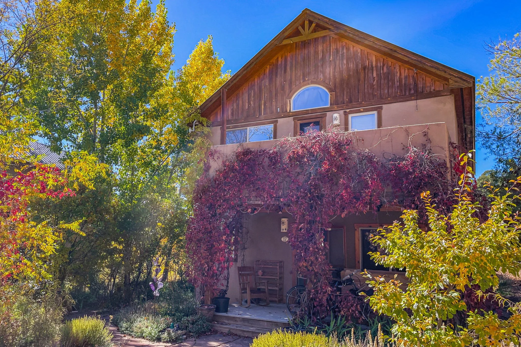 A two-story house with wood siding and a stucco lower level, surrounded by colorful autumn trees and plants, with a covered front porch and a blue sky in the background.