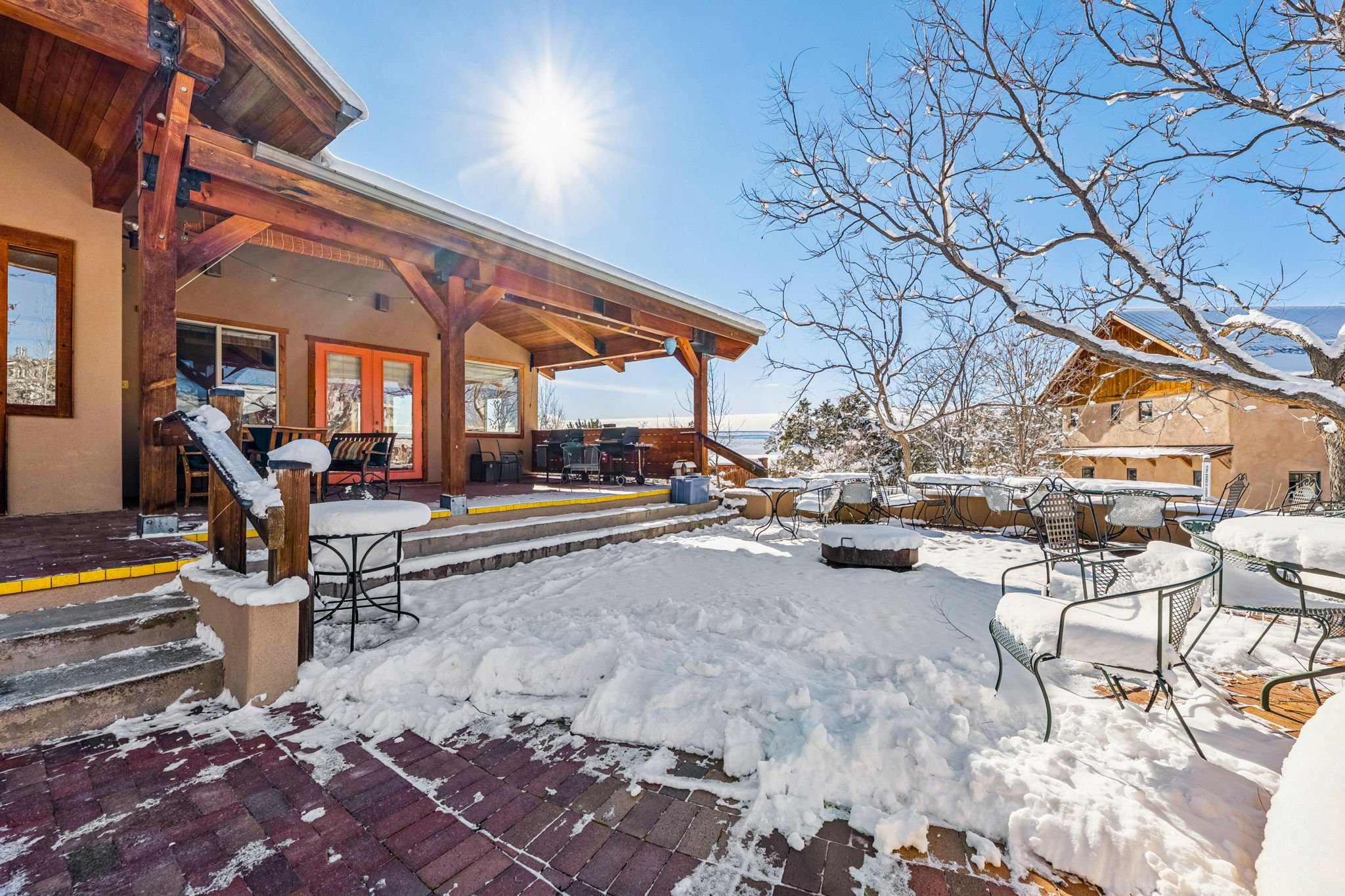 Snow-covered outdoor patio with tables, chairs, and a fire pit in front of a house under a bright sunny sky.