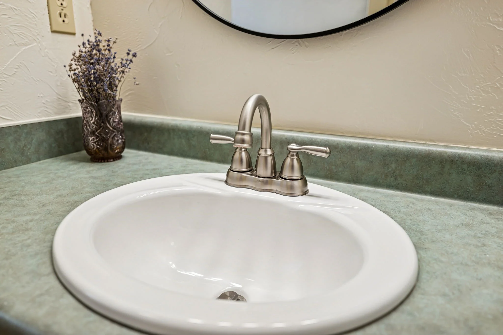 Bathroom countertop with a white sink and a bronze faucet, a decorative vase with dried flowers, mirror, and a wall outlet.