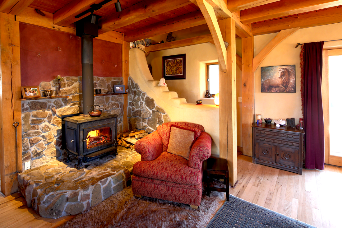 Living room with a stone fireplace, red armchair with a cushion, wooden beams, a window with a ledge, and a small cabinet with decorative items.