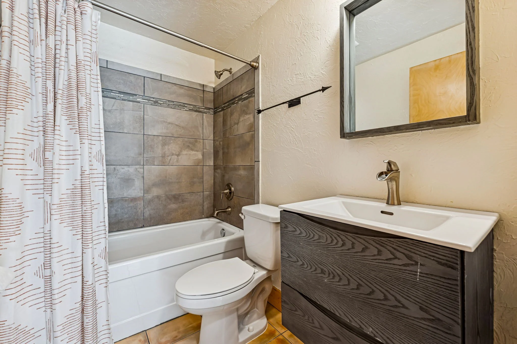 Bathroom with bathtub, toilet, sink, and mirror, beige textured walls, tiled shower area, wood-look flooring.