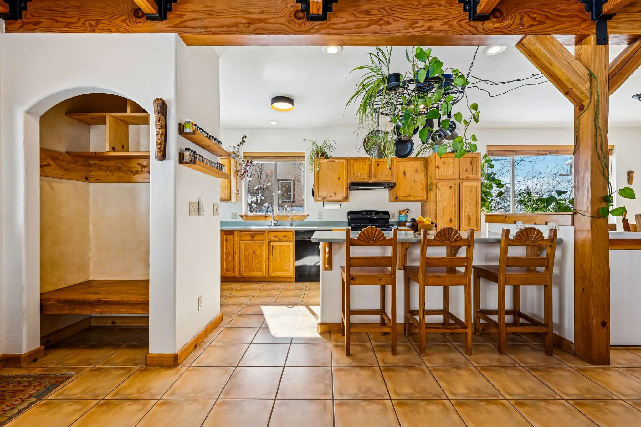 Kitchen with wooden cabinets, tile flooring, and a breakfast bar with four wooden chairs. There are large windows, hanging plants, and kitchen appliances.