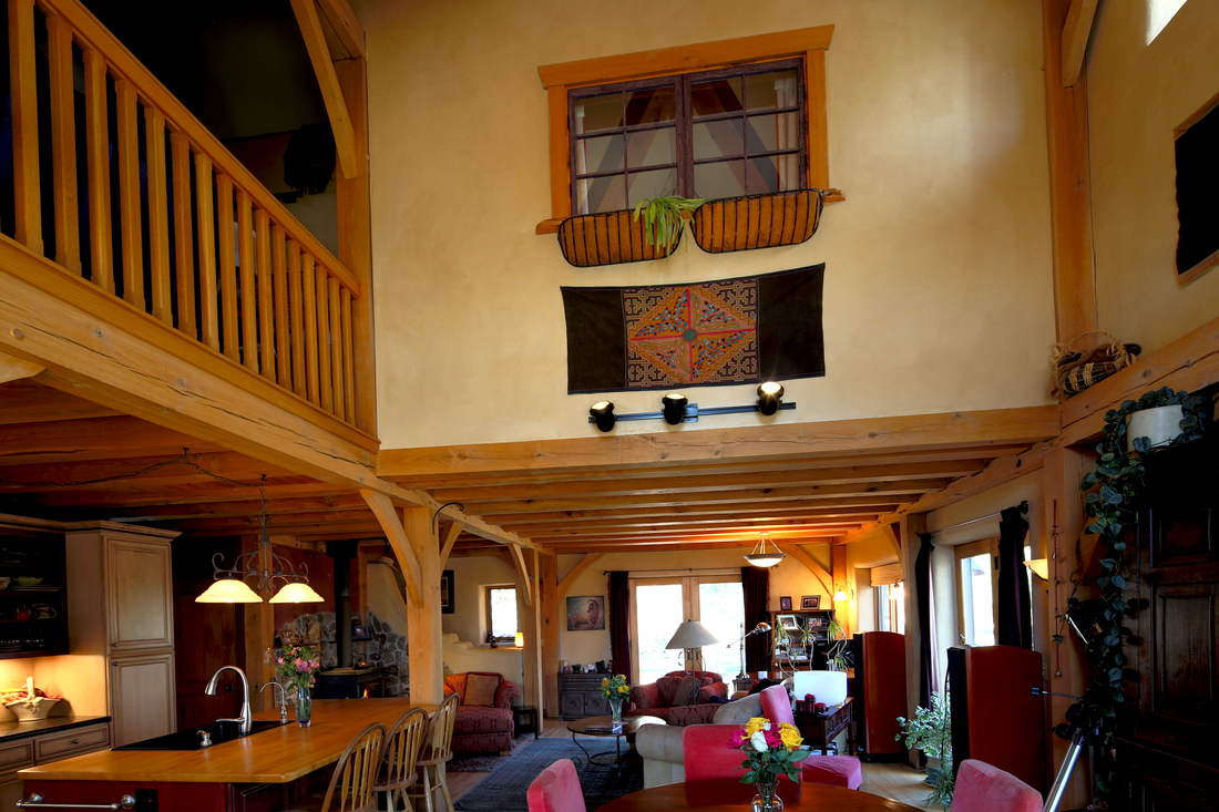 Interior of a cozy, rustic living room with a wooden ceiling and beams, decorated with plants, a tapestry, and furniture including a dining table, chairs, sofas, and lamps.