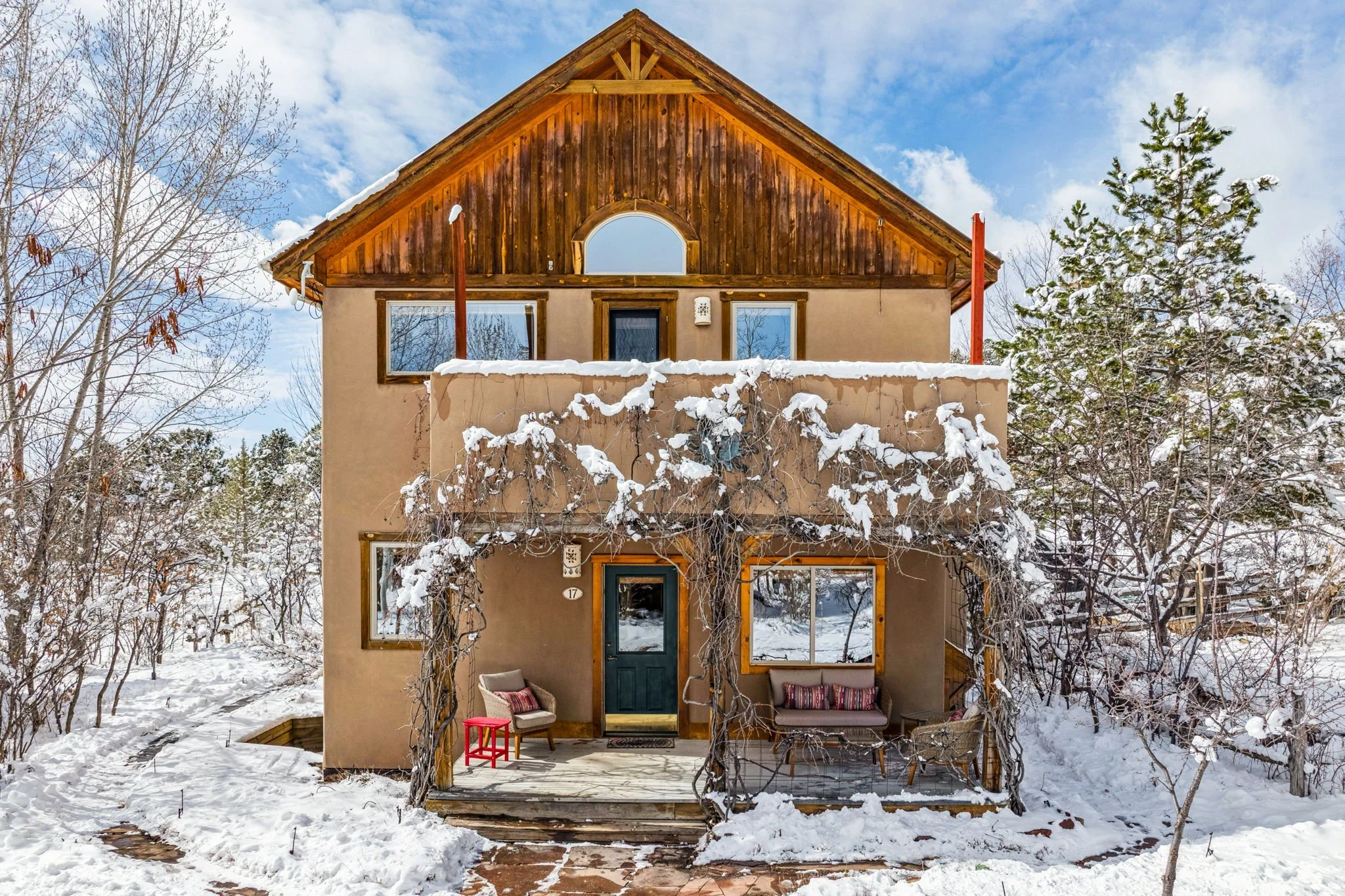 A two-story house with a stucco exterior, wooden accents, and a snow-covered front porch surrounded by trees and snow.