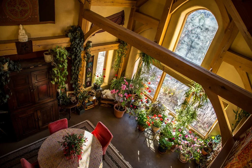 An interior view of a room with a sloped wooden ceiling, decorated with numerous potted plants and flowers near large windows, a round dining table with a floral tablecloth, and a dark wooden cabinet.