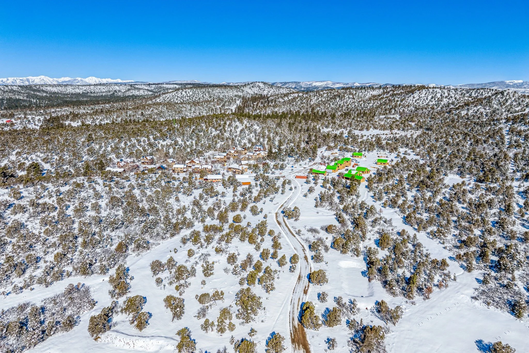 A snow-covered mountainous landscape with a small village, trees, and roads, under a clear blue sky.