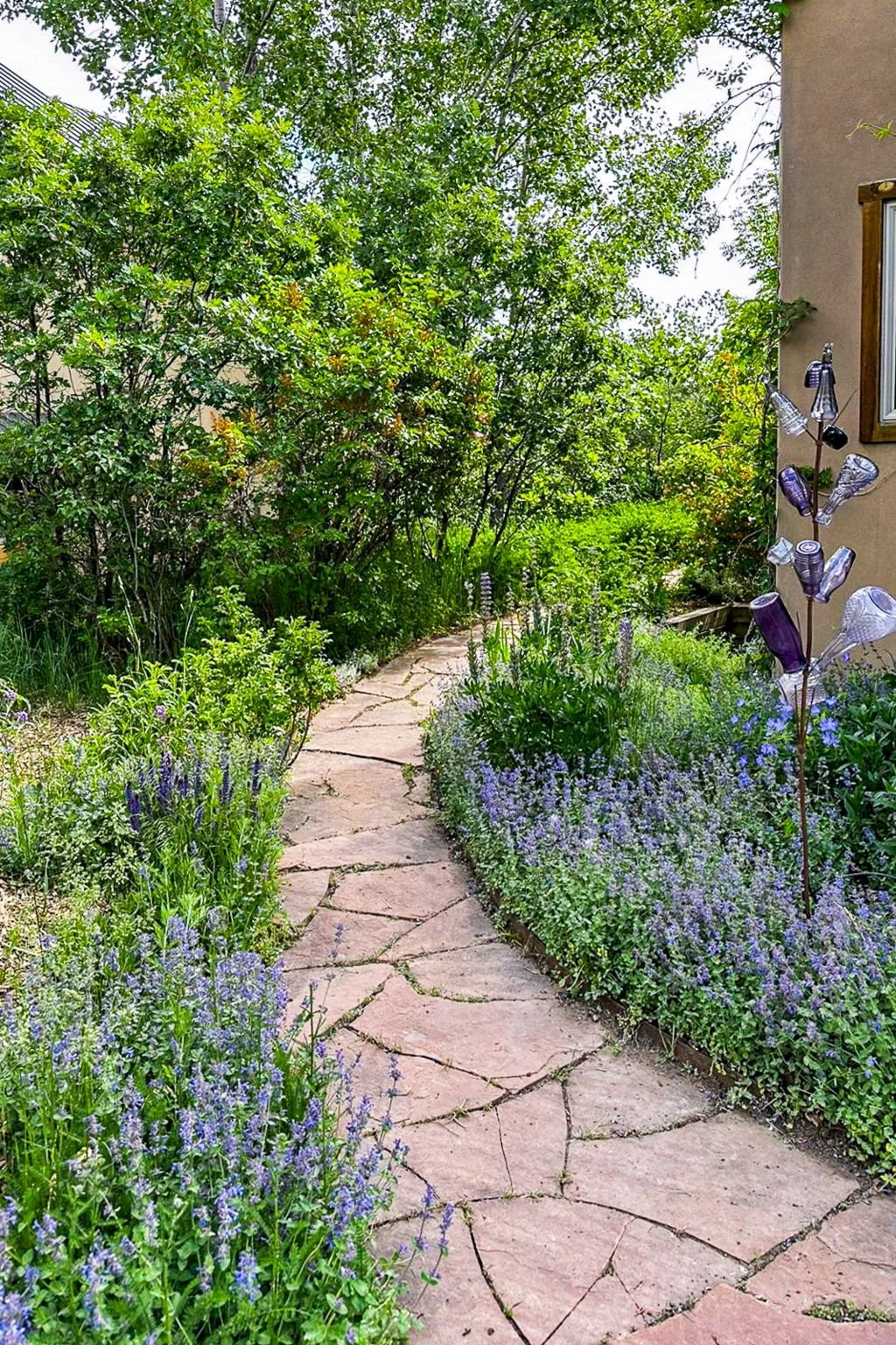 A stone pathway winds through a lush garden filled with purple, blue, and green plants, with dense green trees and shrubs in the background, and a part of a house with wind chimes visible on the right side.
