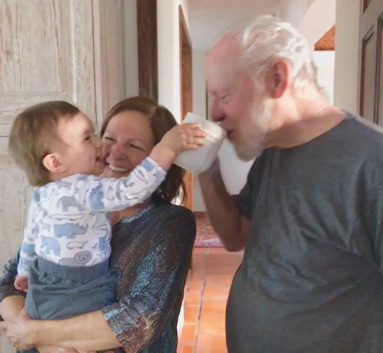 A young boy with a shirt featuring elephants gives a drink to an elderly man with white hair and a beard, while a woman holds the boy and smiles in a cozy home interior.