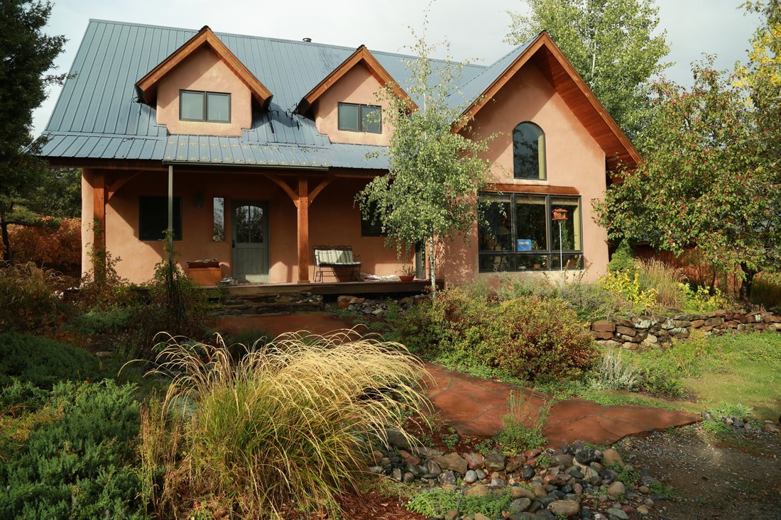 A house with a blue metal roof and pinkish exterior walls, surrounded by trees and plants, with a front porch and a garden.