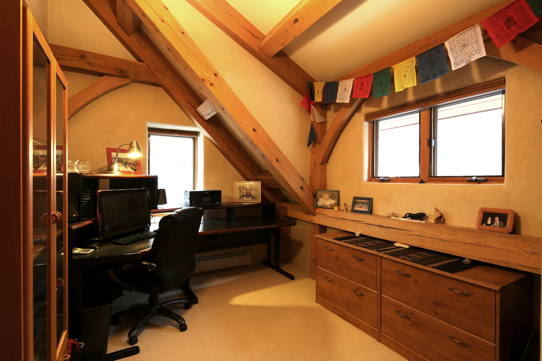 A home office in an attic with wooden beams, a desk with a computer, and framed photographs on a windowsill, decorated with colorful prayer flags.