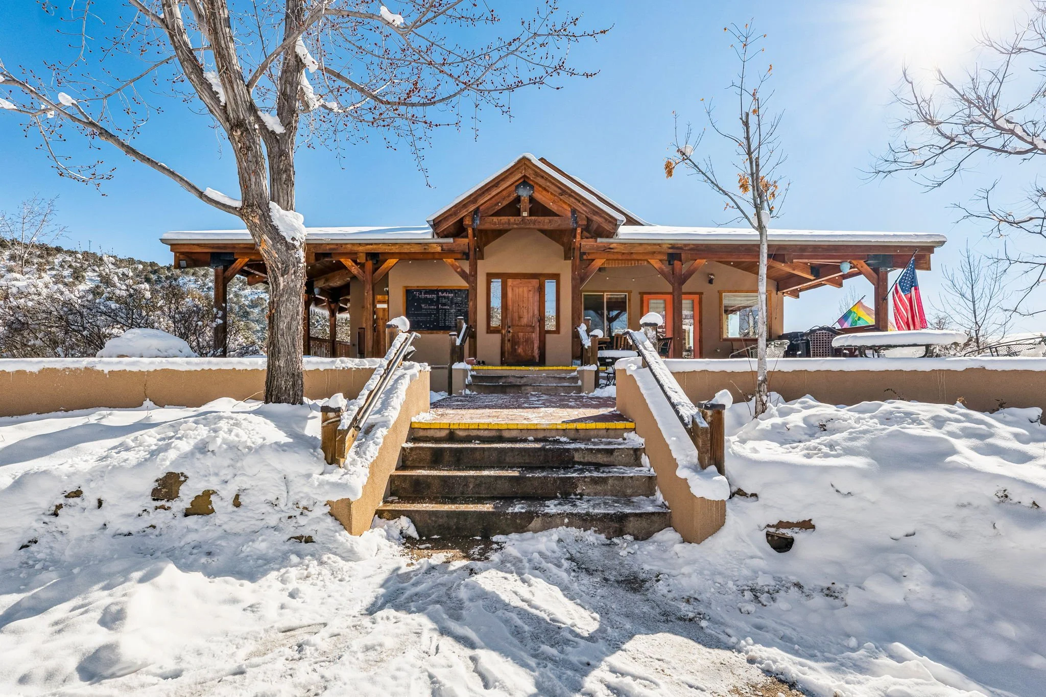 A snow-covered house with a wooden porch, two staircases leading up to the entrance, flanked by snow-covered trees, on a bright sunny winter day.