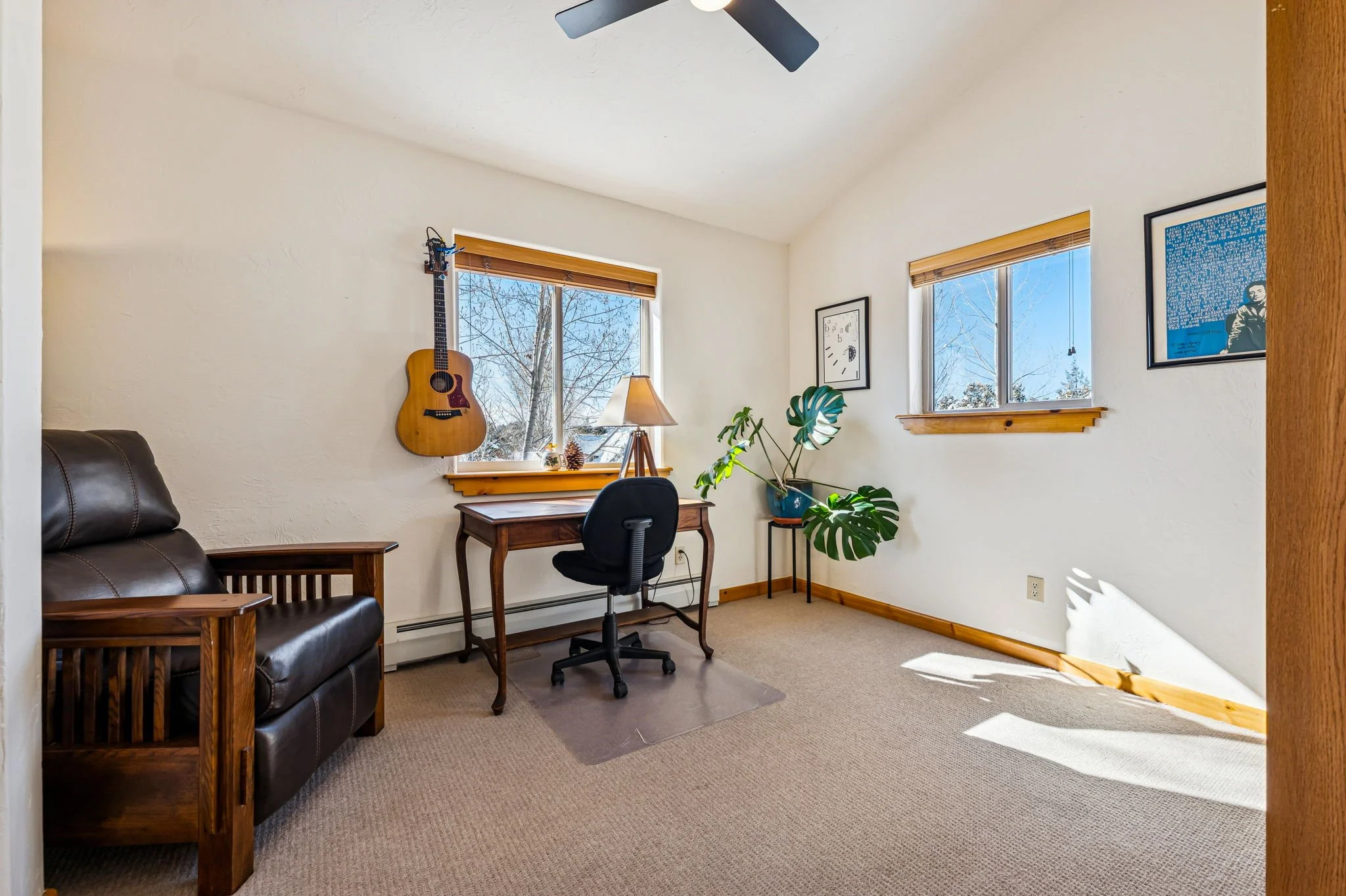 A cozy home office with two windows showing a snowy outdoor scene, a black office chair at a wooden desk, a guitar hanging on the wall, a lamp on the desk, a large potted monstera plant, framed art on the walls, and carpeted flooring with wooden trim