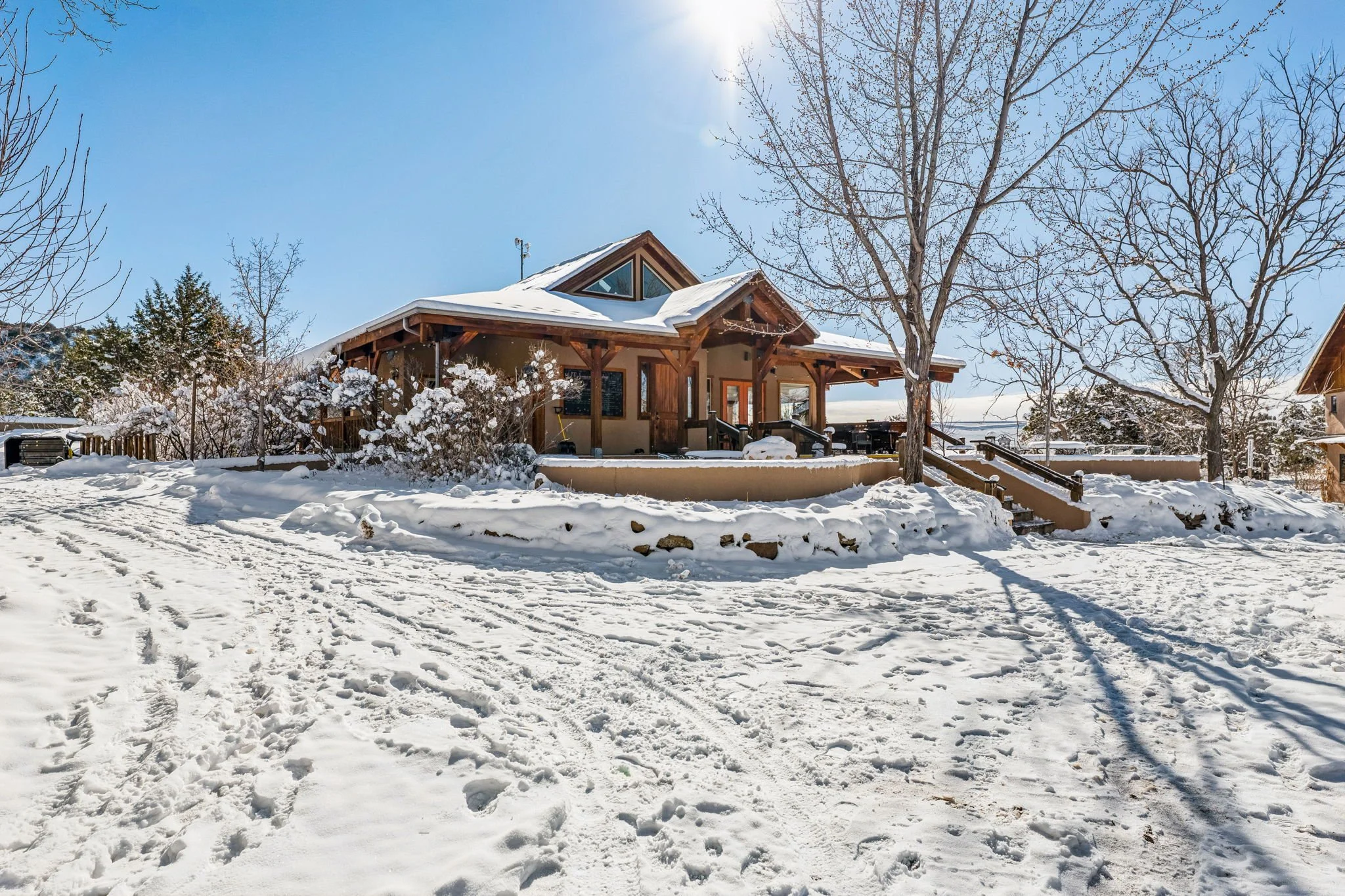 A wooden house with a snow-covered yard and trees, bright sunny sky, and a clear view of the house's porch and steps leading up to the entrance.
