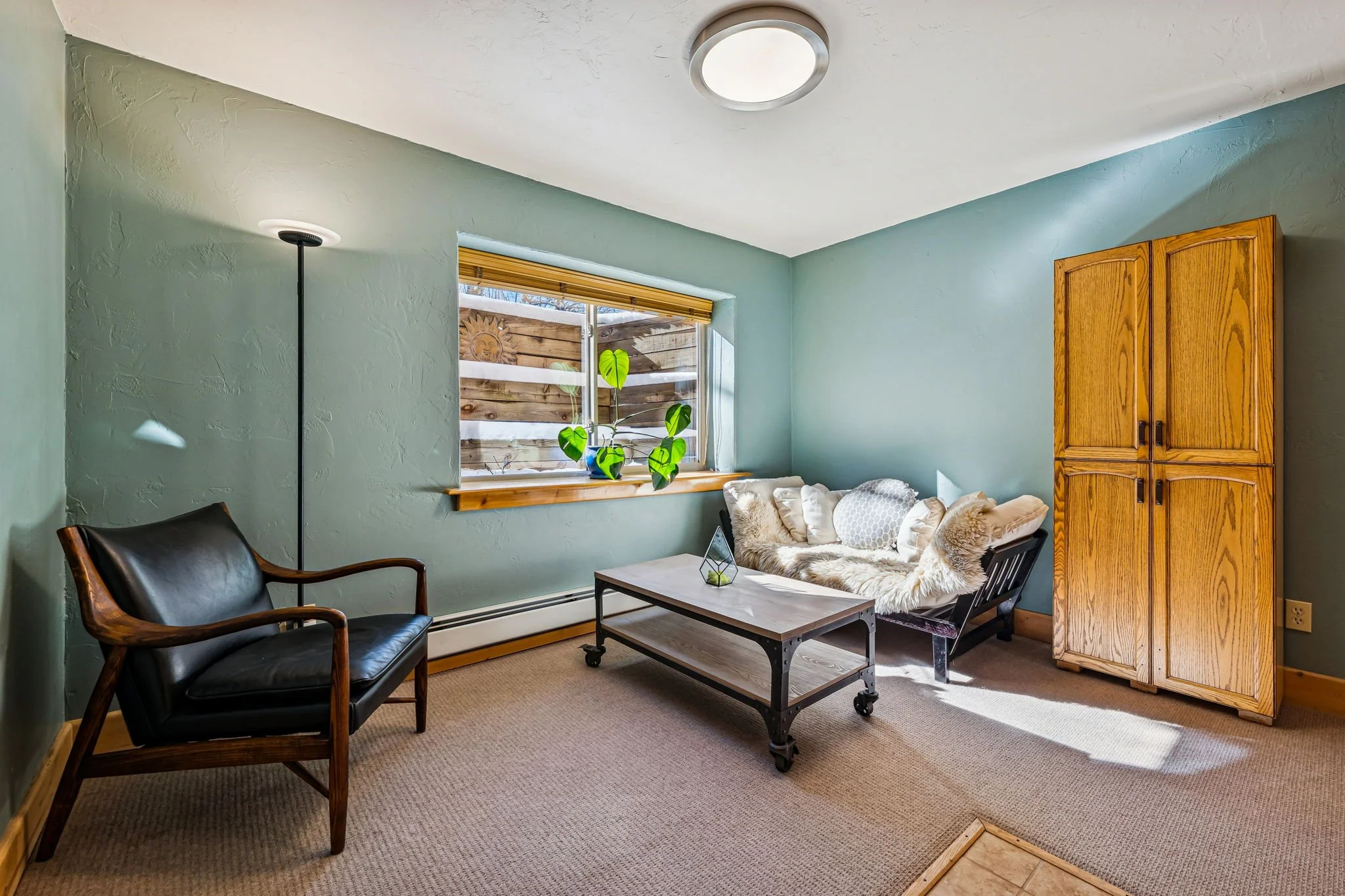 Living room with light green walls, a window with wooden trim and blinds, a black leather armchair with wooden frame, a black futon with plush cushions, a wooden cabinet, a black metal coffee table, and a beige carpet.