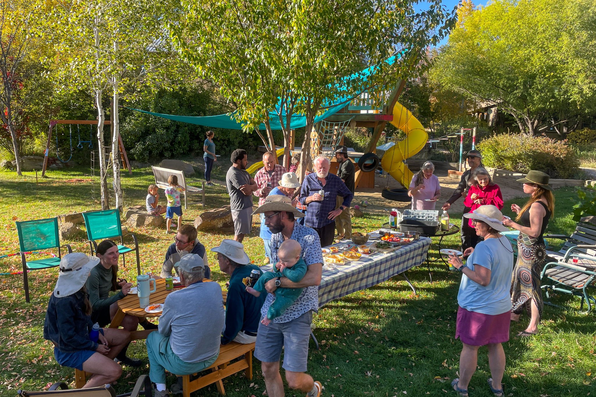 A backyard gathering with people enjoying a sunny day, some sitting at a table, others standing and chatting, in front of a children's play area with a slide and swings, surrounded by trees and greenery.