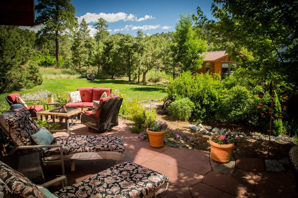 A backyard patio with outdoor furniture, including floral and red cushions, surrounded by potted plants, trees, and a lush green yard, with a wooden shed in the background and a mountain view under a partly cloudy sky.