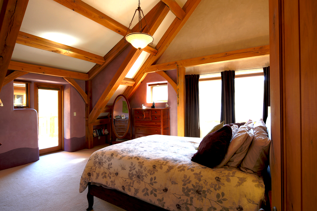 Cozy bedroom with sloped ceiling, wooden beams, a bed with floral bedding, and multiple windows with black curtains.