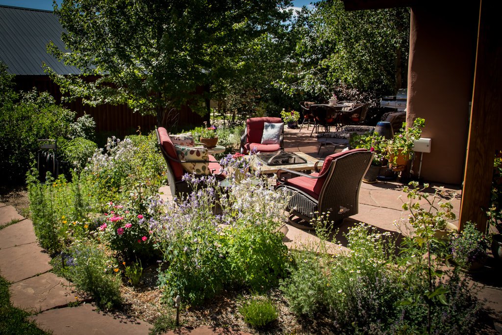 A backyard patio area with garden beds of various flowers and plants, outdoor seating with cushioned chairs and a fire pit, surrounded by trees and greenery.