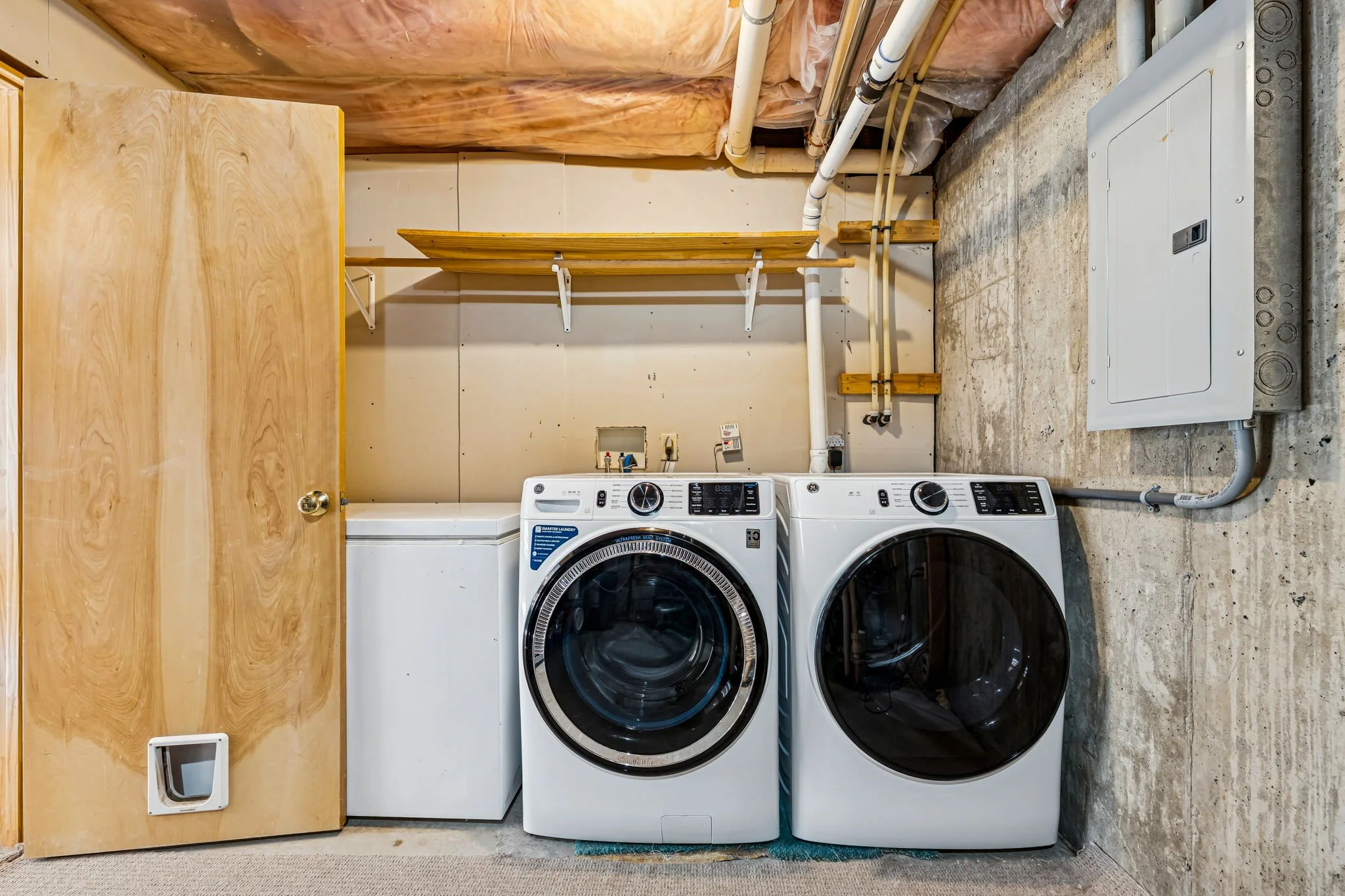 Laundry room with front-loading washer and dryer, a white chest freezer, wooden door, wall-mounted shelves, exposed pipes, and a concrete floor.