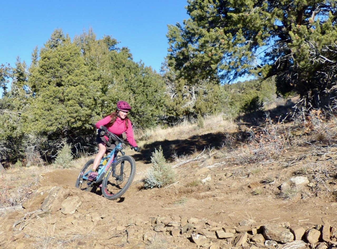 A young girl wearing a pink helmet and shirt riding a mountain bike on a dirt trail surrounded by shrubs and trees in a sunny outdoor setting.