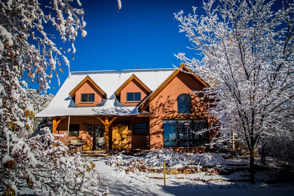A house with a snow-covered roof and surrounding snow-covered trees under a clear blue sky.