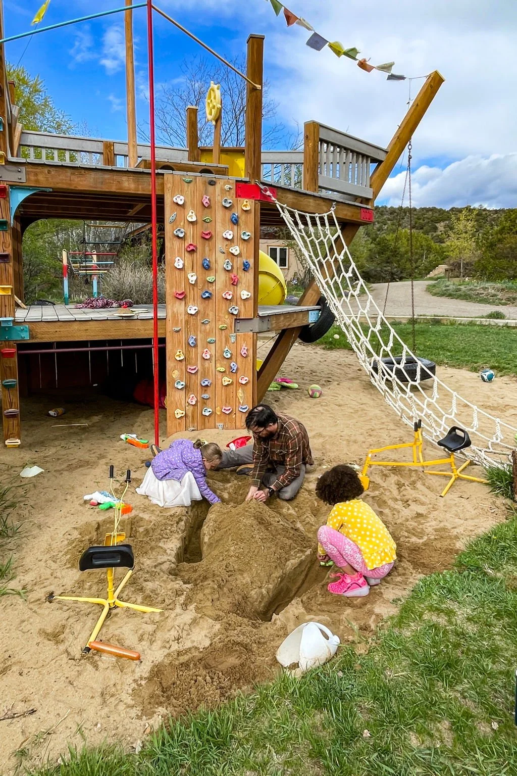 Children and an adult working together in a sandbox under a wooden play structure with a rock climbing wall, slide, and swings, in a park with grass and trees.