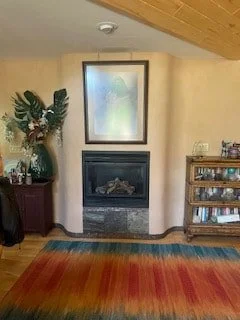 Living room with a fireplace, large mirror above it, a floral arrangement on a stand to the left, and a wooden shelving unit with books and decor to the right. Rug with red, orange, and yellow stripes on the floor.