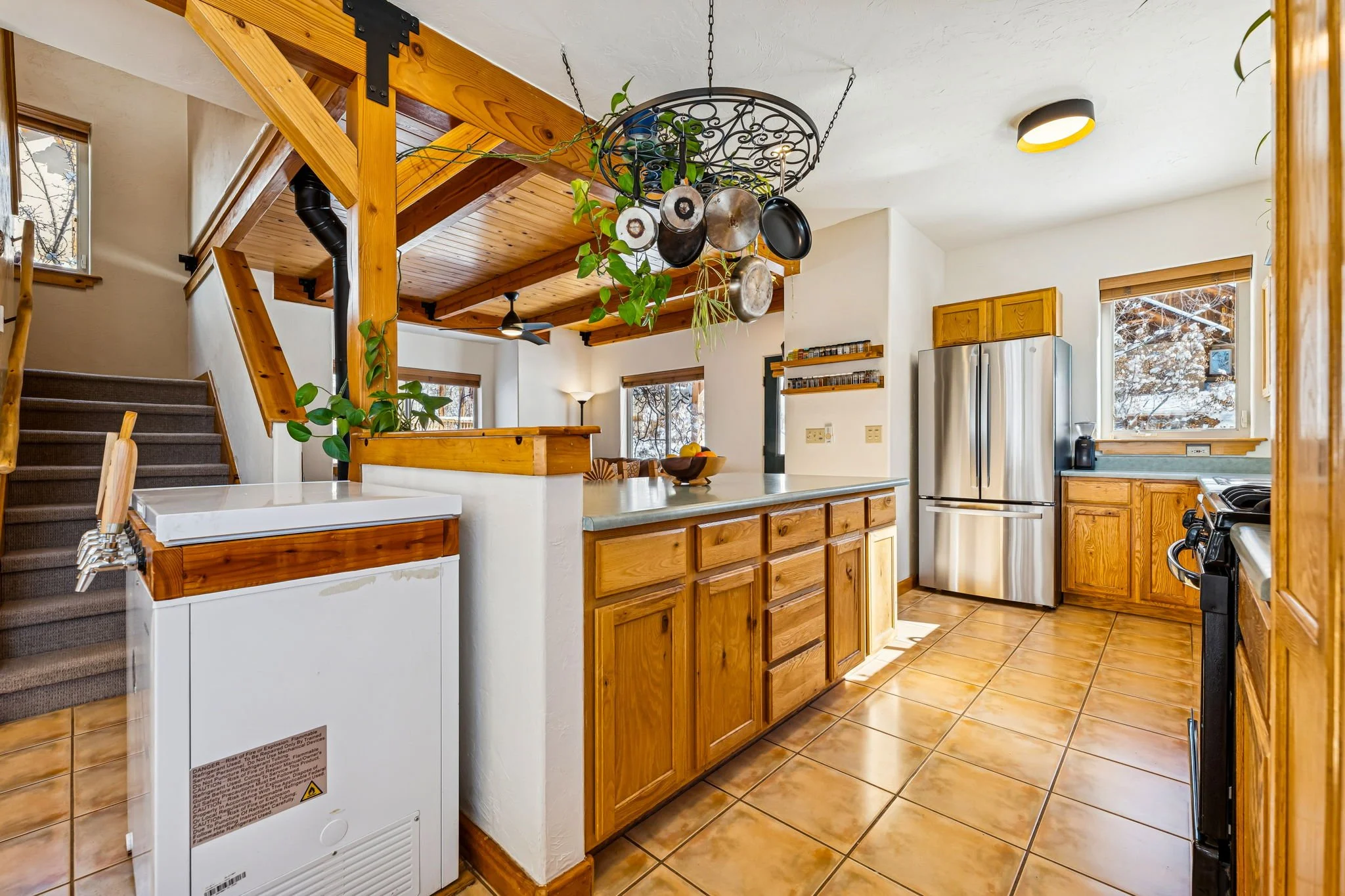 Kitchen with wooden cabinets, stainless steel refrigerator, and tiled floor, featuring a ceiling-mounted pot rack with hanging pots and pans.