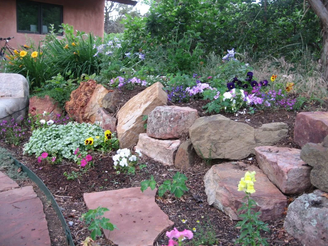 A garden with colorful flowers including pansies, petunias, and snapdragons, surrounded by large stones and rocks, with a house and greenery in the background.