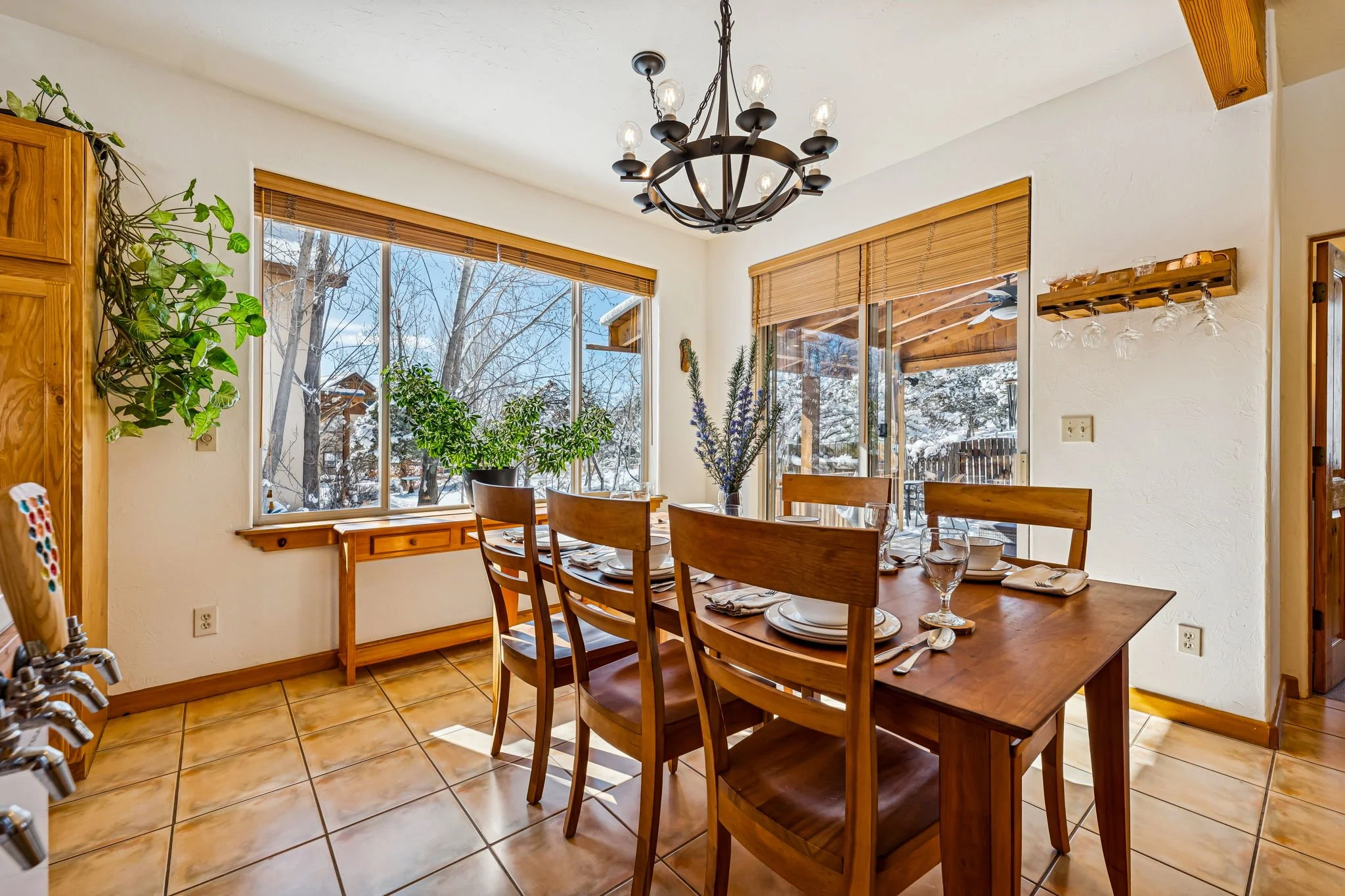 Dining room with a wooden table set for four, large windows showing a snowy outdoor scene, potted plants, a chandelier, and glassware.