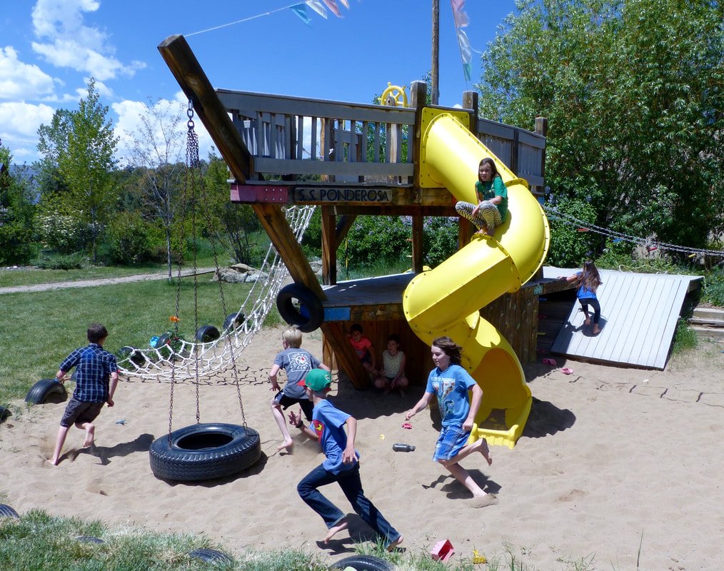 Children playing around a wooden playground structure with a yellow slide and tire swings on a sandy surface under a blue sky.