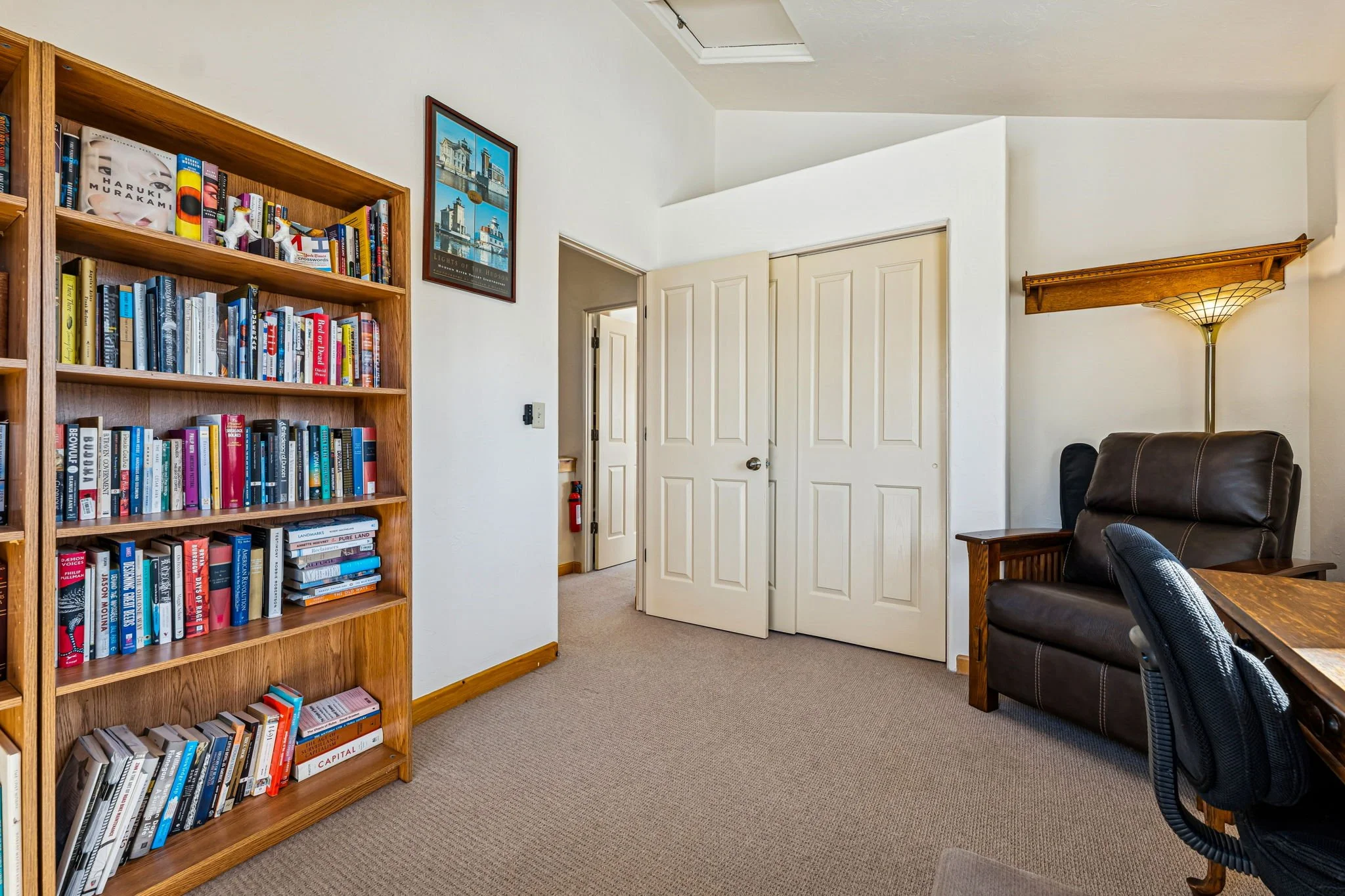 A room with a wooden bookshelf filled with books on the left, a framed picture of architecturally interesting buildings on the wall, an open closet door with double doors in the center, a leather armchair next to a lamp with a wooden shelf, and part 
