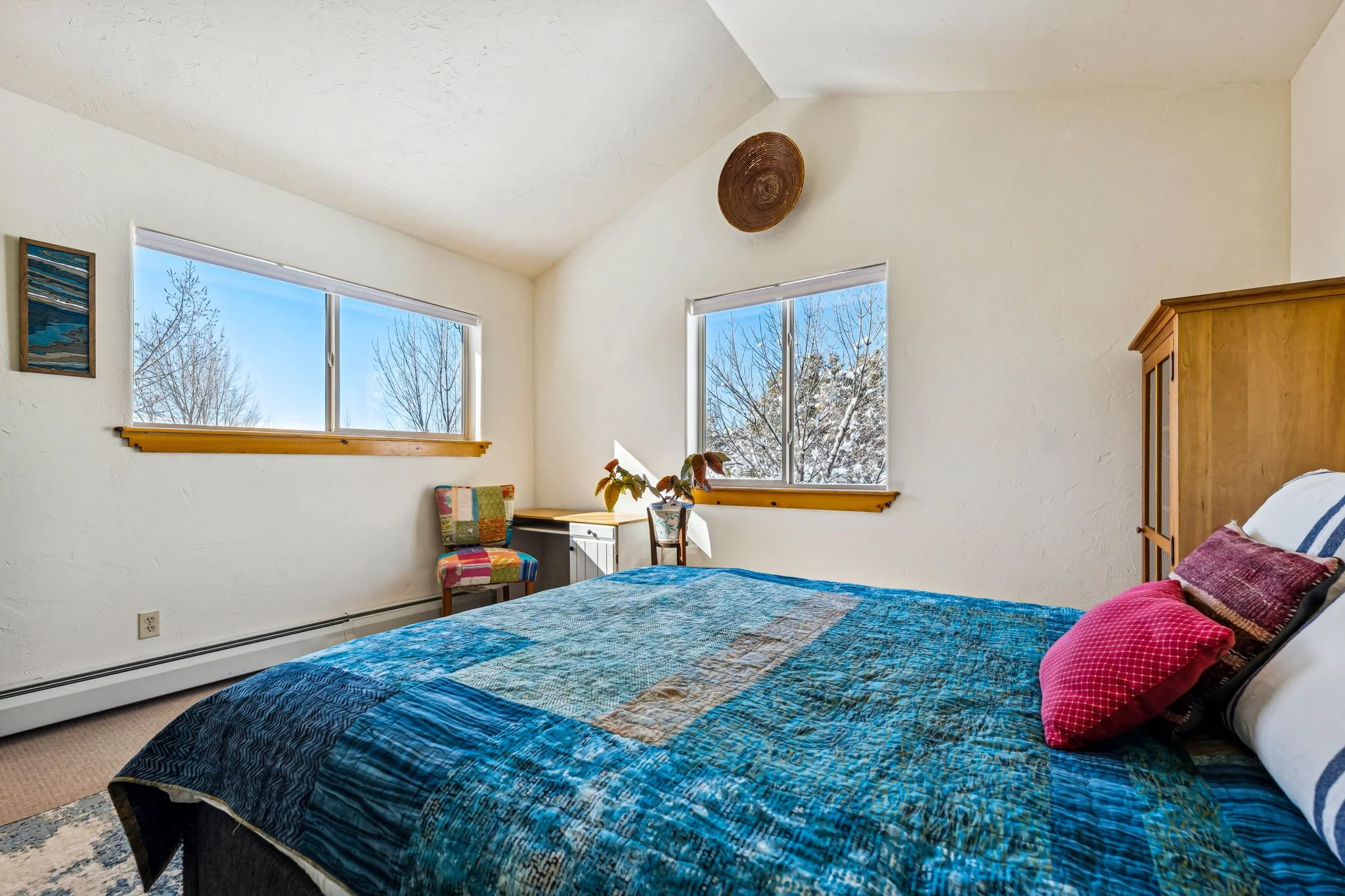 A bedroom with two windows showing snowy trees outside, a wooden headboard, a blue bedspread, red powder and striped pillows, a colorful chair, small dresser, and a potted plant, with a white wall and ceiling.