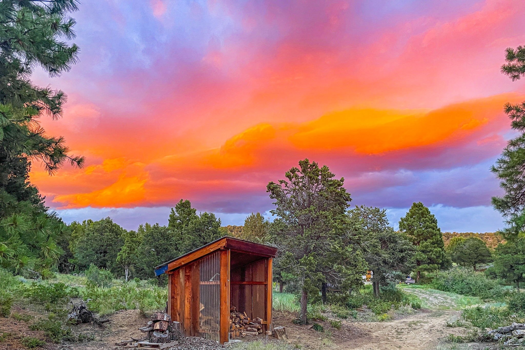 A sunset sky with vibrant pink, orange, and purple clouds over a wooded landscape with a small wooden shelter and a dirt path.